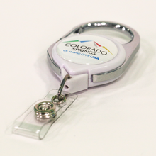 Load image into Gallery viewer, Carabiner badge reel with label that has the full color Colorado Springs: Olympic City USA logo on top of a white background laying flat on a white surfafce
