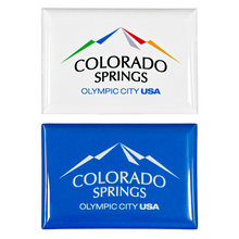 Load image into Gallery viewer, 2 rectangular, shiny magnets. Both have printed artwork. One includes a full colored Colorado Springs: Olympic City USA logo on a white background, and the other is the same logo white on top of a blue background.
