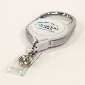 Carabiner badge reel with label that has the full color Colorado Springs: Olympic City USA logo on top of a white background laying flat on a white surfafce