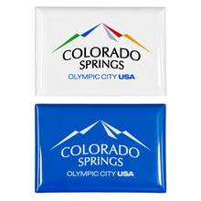 Load image into Gallery viewer, 2 rectangular, shiny magnets. Both have printed artwork. One includes a full colored Colorado Springs: Olympic City USA logo on a white background, and the other is the same logo white on top of a blue background.  
