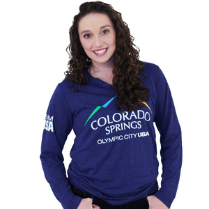 Model wearing dark blue long sleeve v-neck shirt. Shirt has full color logo for the city of Colorado Springs: Olympic City USA printed on front. Team USA printed in white on the left sleeve on the upper arm.