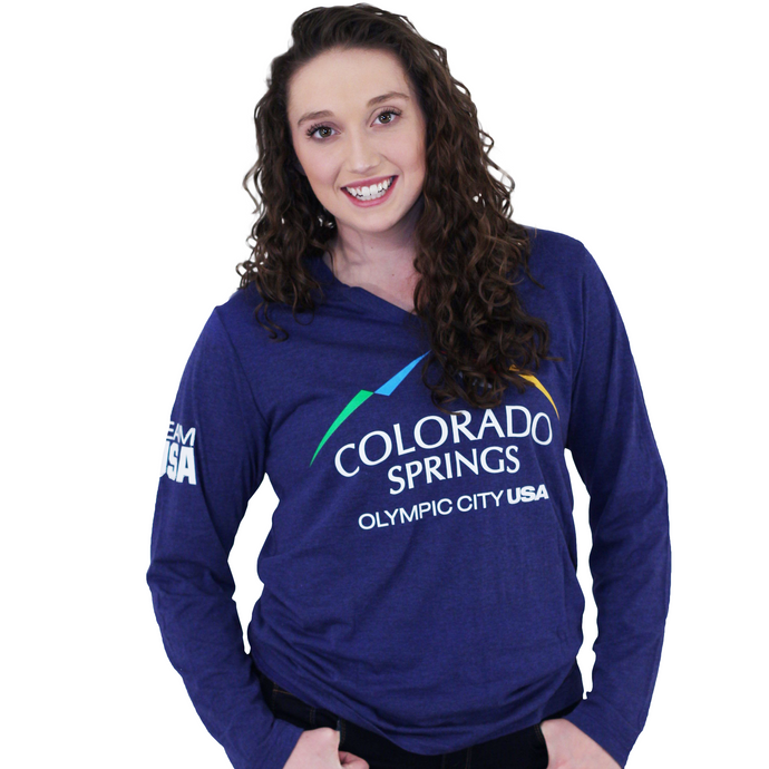Model wearing dark blue long sleeve v-neck shirt. Shirt has full color logo for the city of Colorado Springs: Olympic City USA printed on front. Team USA printed in white on the left sleeve on the upper arm.